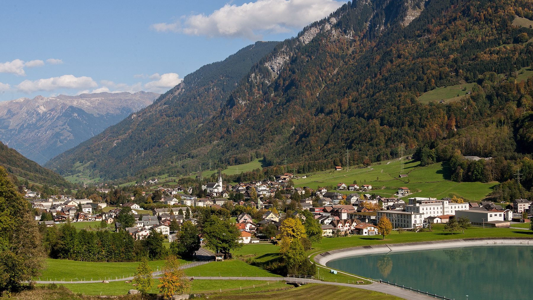 Blick über Linthal im Herbst: das Dorf im Talboden mit Kirchturm, dahinter
              bewaldete Bergflanken, im Vordergrund Wiesen und das Wasserbecken des
              Kraftwerks.